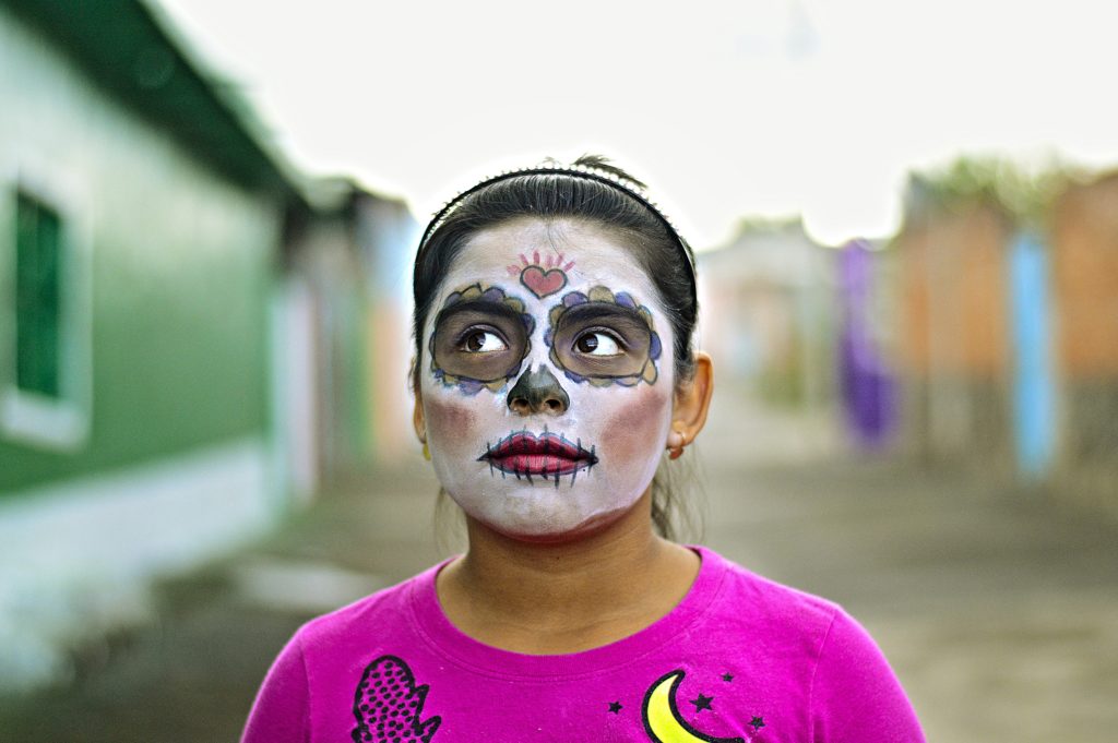 one of our favorite group trip ideas is to visit Mexico for dia de los muertos. photo of a young woman wearing traditional face paint with a pink shirt and blurred background