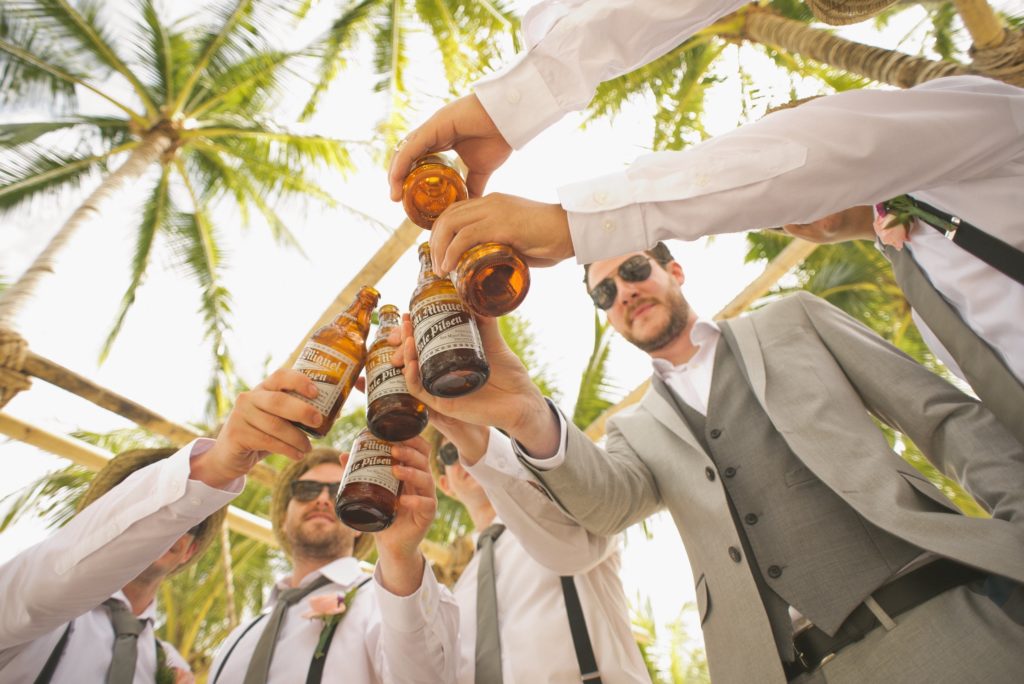 group vacations palm trees in the background looking up at a group of groomsmen and a groom cheers'ing with beers