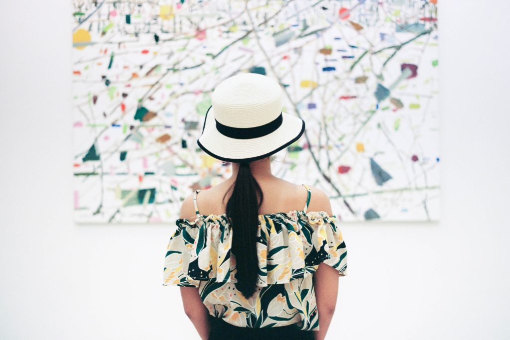 girl with a black ponytail, colorful ruffled shirt and white and black floppy hat from behind stands looking at an abstract colorful painting against a white background