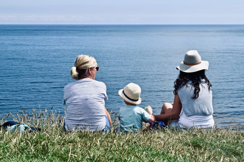 a grandmother, her daughter, and her grandson sit on grass overlooking the ocean as fun things to do on vacations for multigenerational families