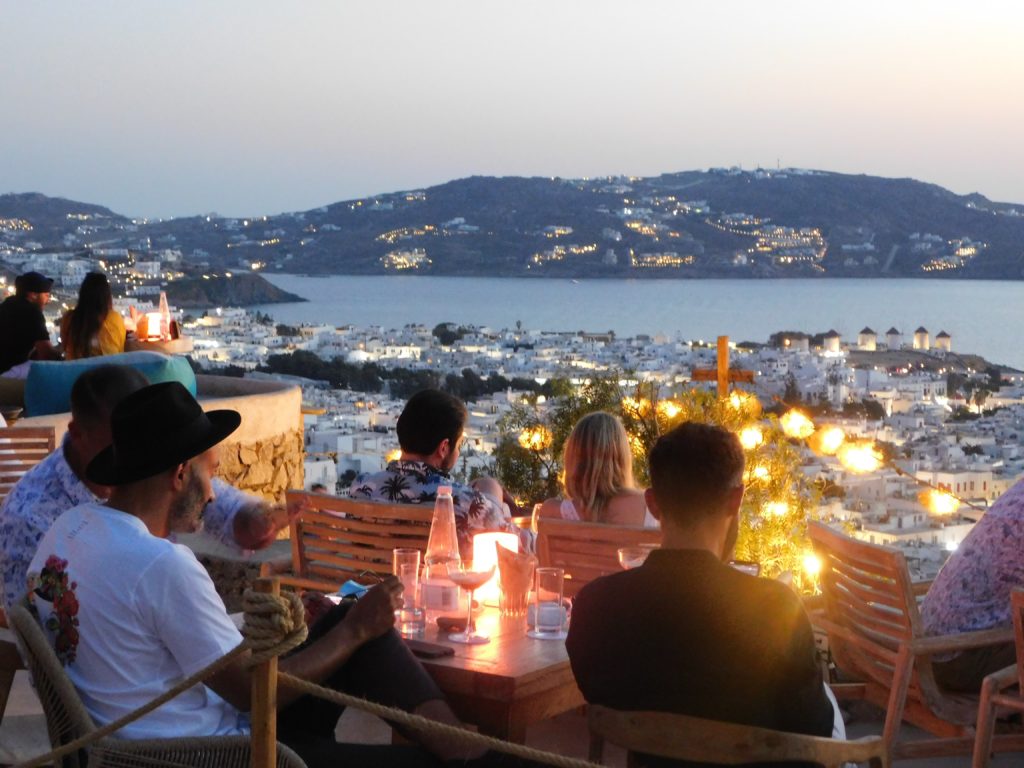 friends sit at tables looking out over a Greek island full of white buildings with the other side of the island over the sea in the distance at sunset