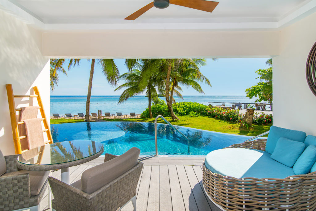 view looking out from the room with table and chairs, wicker couch with blue cushions and direct access into private pool with ocean and palm trees behind  