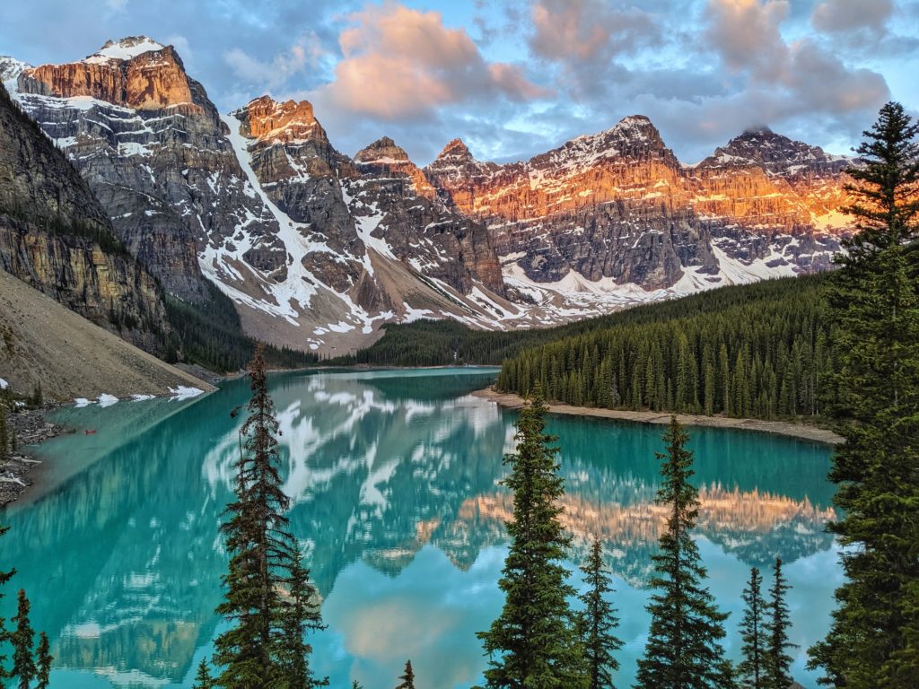 Mountains with snow in the background and bright blue lake with mountains and pine trees reflecting in the water