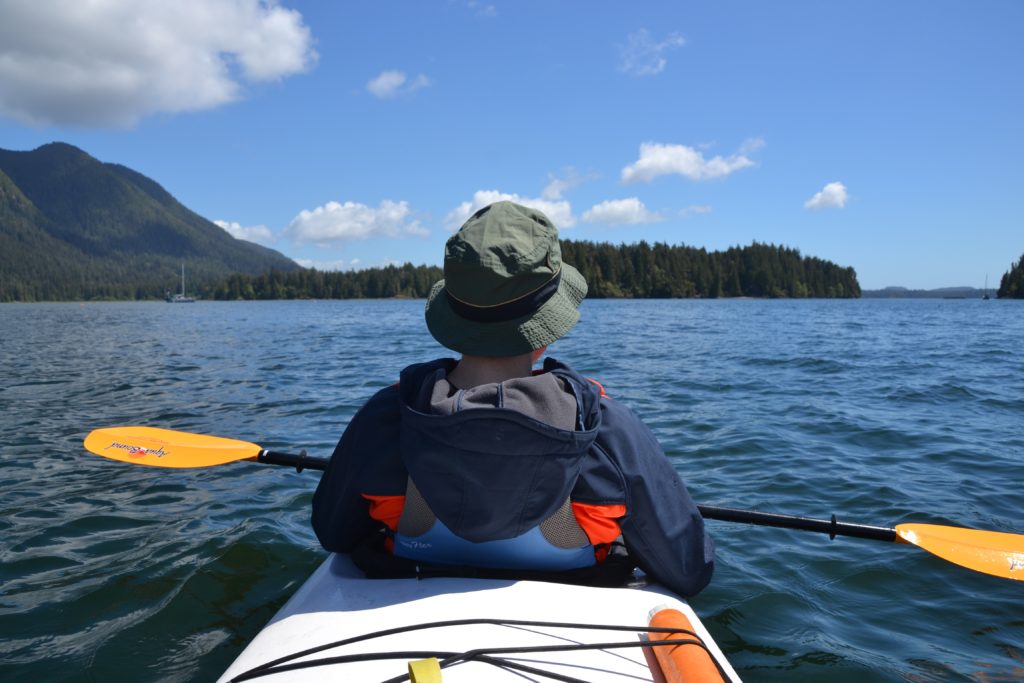 man sits in kayak wearing bucket hat with green mountains in background 