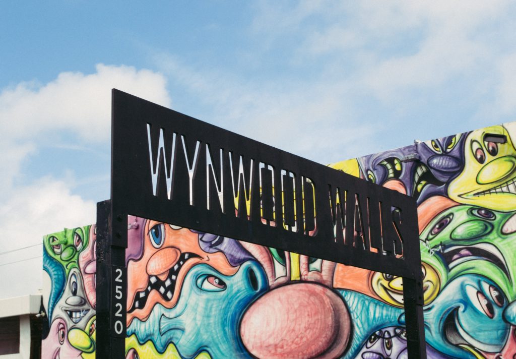 wynwood walls written through steel cutout with graffiti mural in the background of colorful funky looking faces