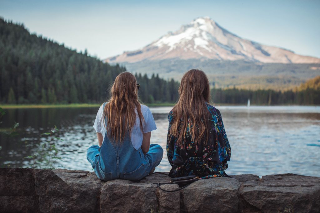 two girls on a girls trip in the mountains with their back to the camera sitting on a rock wall with a lake and mountain in the background