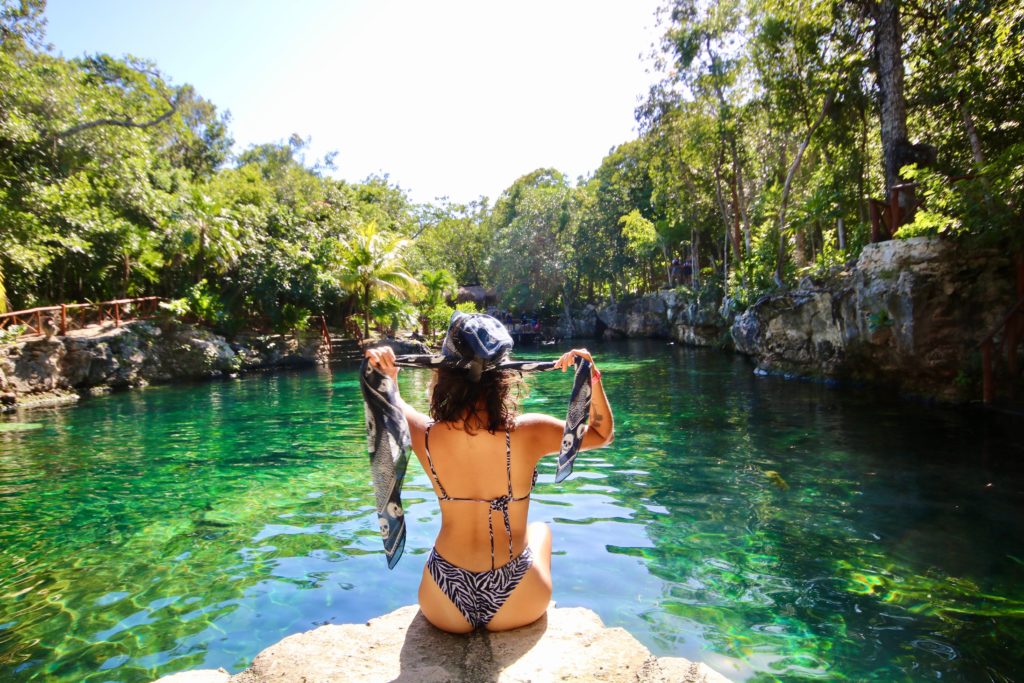 a girl sits on a ledge overlooking a cenote full of emerald green waters 