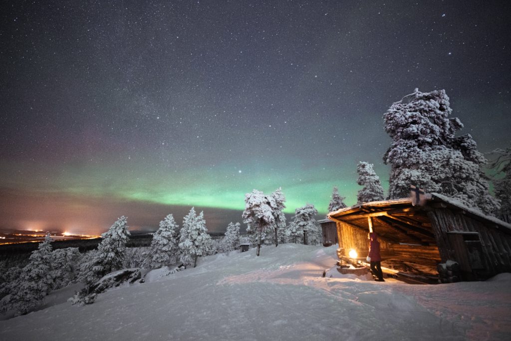 a cabin with a person standing out front looking up at the northern lights in the sky over snow covered trees.