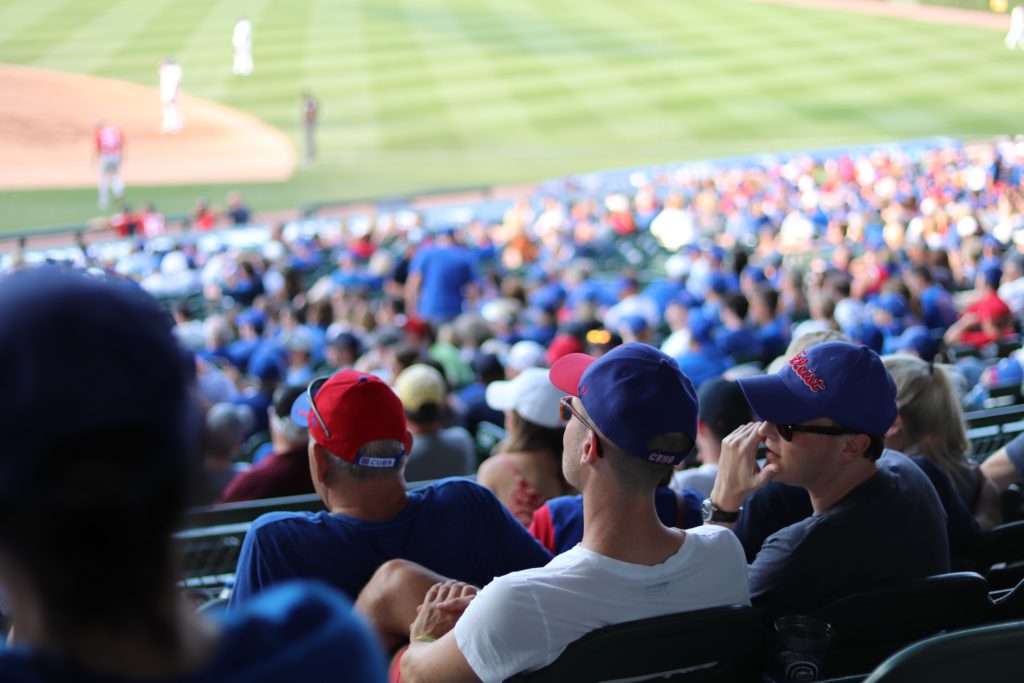 shot of the crowd at Wrigley Field. Blurred heads in the stands in the background and 2 guys wearing Cubs gear in the foreground.