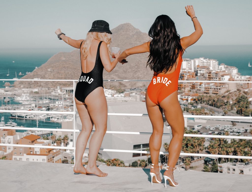 2 girls dance in one-pieces overlooking downtown Cabo. One says Bride on the back and the other says Squad.