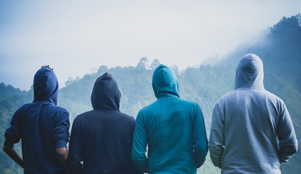 four men standing looking away from the camera at tree-covered mountains with sweatshirts and hoods on